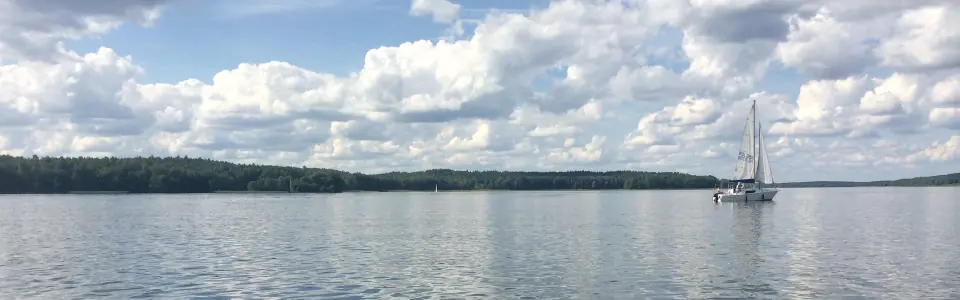 Jacht pod żaglami na Jeziorze Charzykowskim, nad lasem na drugim brzegu cumulusy