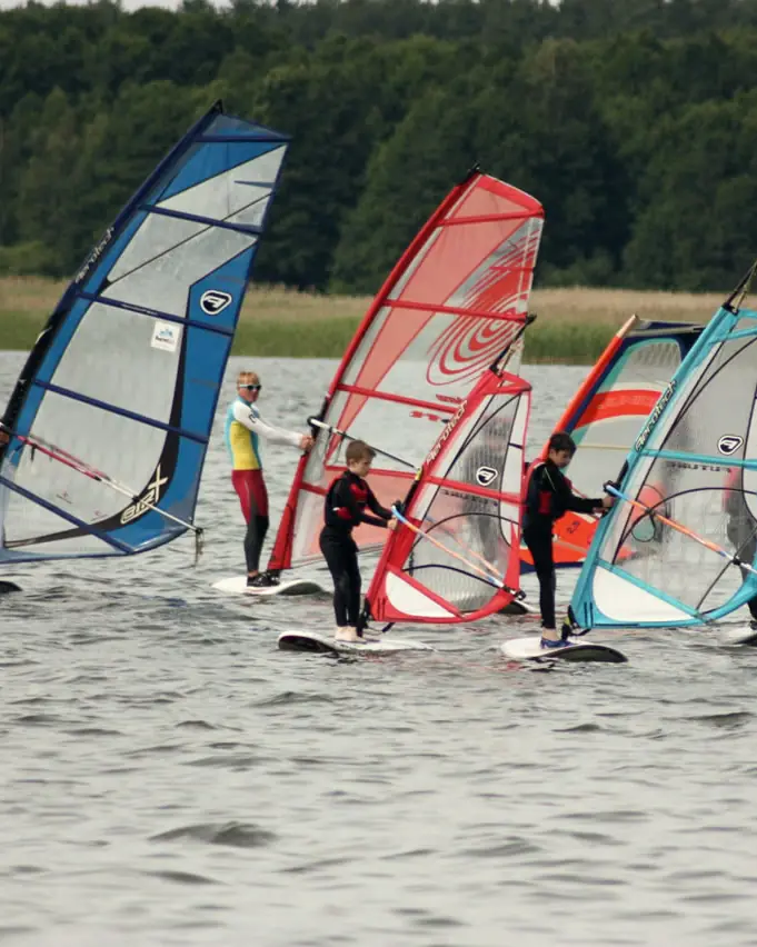 Grupa uczniów na szkolnych deskach z kolorowymi żaglami podczas lekcji windsurfingu
