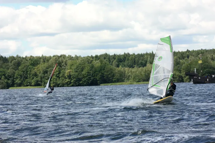 Dwóch windsurferów w ślizgu na wzburzonym jeziorze, las na drugim brzegu